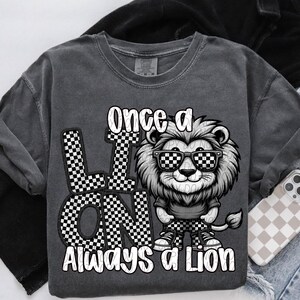 May include: A gray t-shirt with a black and white checkered design and a cartoon lion wearing sunglasses. The text "Once a Lion Always a Lion" is printed on the shirt.