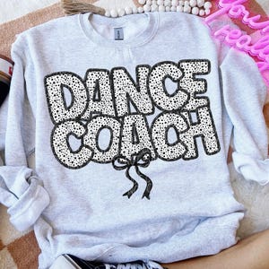 May include: A grey sweatshirt with the text "DANCE COACH" in black outlined letters with a white polka dot pattern. A black bow is below the text.