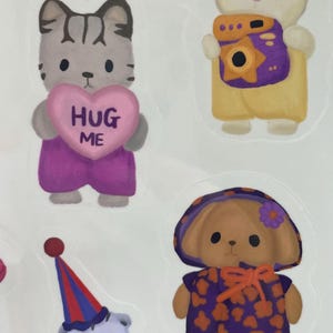 May include: A sheet of colorful stickers featuring cartoon animals. Includes a cat holding a heart that says "HUG ME", a bunny with a camera, a dog in a hat, and a clown-like character. The stickers are in various colors.