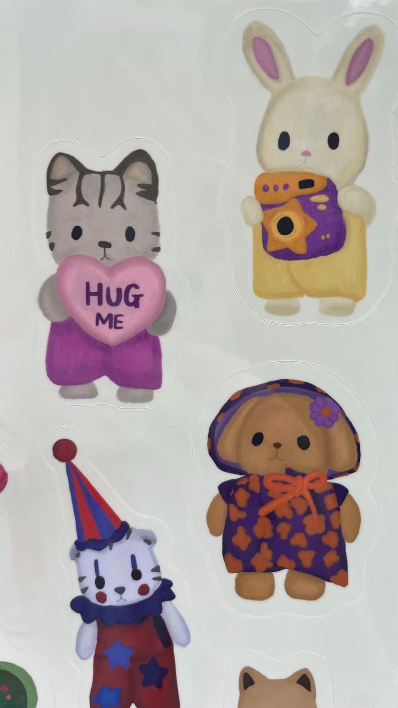 May include: A sheet of colorful stickers featuring cartoon animals. Includes a cat holding a heart that says "HUG ME", a bunny with a camera, a dog in a hat, and a clown-like character. The stickers are in various colors.