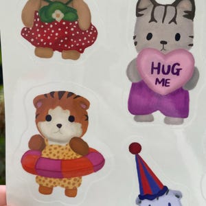 May include: A sheet of colorful stickers featuring cartoon animals. The stickers include a bear in a red dress, a cat holding a pink heart with "HUG ME" on it, a tiger with a swim ring, a clown, and a bunny.