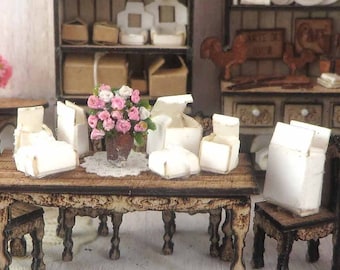 May include: A miniature dollhouse kitchen set with a wooden table and chairs, two wooden cabinets with shelves, and a wooden counter with drawers. The table is set with white boxes and a vase of pink flowers. The cabinets are filled with miniature dishes and other kitchen items. The counter has a small bowl of fruit on top.