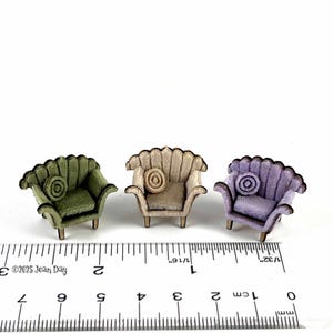 May include: Three miniature armchairs in olive green, beige, and lavender. Each chair has a scalloped back, a round cushion, and wooden legs. The chairs are approximately 2.5 cm in height, with a ruler in the foreground.