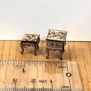 May include: Two wooden miniature tables with intricate floral designs. The larger table measures 1 3/32 inches tall and the smaller table measures 1 1/16 inches tall.