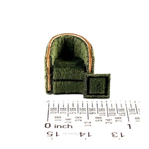 Puede incluir: Miniatura de sillón verde con una mesa auxiliar cuadrada a juego. La silla tiene respaldo y reposabrazos redondeados, con un ribete marrón claro. El conjunto está hecho de un material difuso y texturizado. La escala está indicada por una regla en pulgadas.