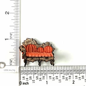 Puede incluir: Un broche decorativo en miniatura con forma de sofá, con un marco marrón oscuro ornamentado con detalles de hojas y cojines naranjas en forma de calabaza. El objeto está junto a una regla que muestra un ancho de 3,8 cm y 2,5 cm de alto.