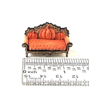 May include: A miniature sofa with a dark wood frame and orange textured upholstery, featuring three orange pumpkin-shaped cushions. A ruler placed in front shows the sofa is approximately 1.25 inches wide and 1 inch tall at the highest point.