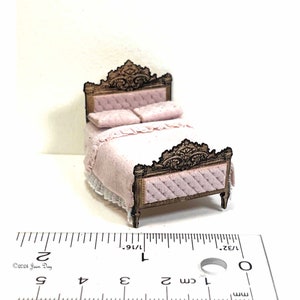 May include: A miniature dollhouse bed with a carved wooden headboard and footboard. The bed is made up with a pink quilted bedspread and two pillows. The bed is approximately 1 inch wide.