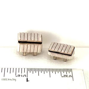 May include: Two miniature white upholstered ottomans with black trim and wooden legs. The ottomans are sitting on a white surface with a ruler in the foreground. The ruler is marked in inches and shows 1/16th increments.