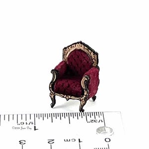 May include: A miniature red velvet armchair with gold accents and ornate detailing. The chair is approximately 1 inch tall and is sitting on a ruler.