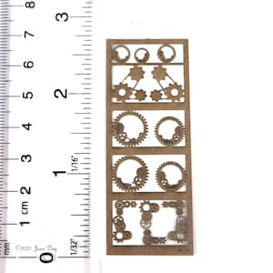 May include: A sheet of brown paper with a ruler for scale. The paper has five rows of cut-out gears in different sizes and shapes.