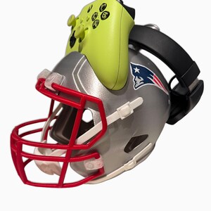 May include: A silver football helmet with a red face mask and the New England Patriots logo. A neon green game controller is attached to the top, and a black headset is on the side. The helmet is a sports-themed item.