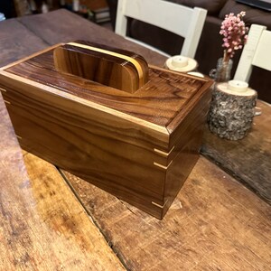 May include: A wooden box with a hinged lid and a handle on top. The box is made of dark wood with light wood accents. The box is sitting on a wooden table.