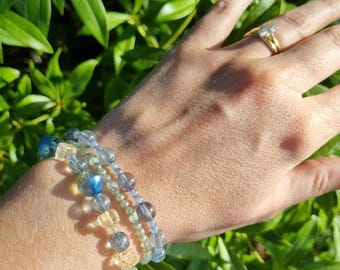 Op de afbeelding: Drie armbanden met kralen in blauwe, gele en heldere tinten zijn op een pols gestapeld. De armbanden hebben verschillende kralenvormen en -maten. Een gouden ring met een grote steen is ook zichtbaar. De achtergrond is groen gebladerte.