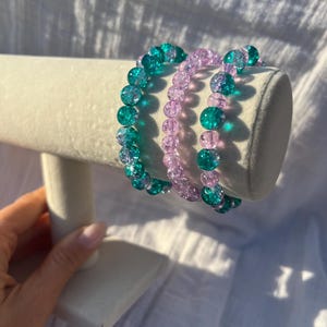 May include: Three beaded bracelets on a white stand. The bracelets have translucent, faceted beads in teal and lavender. The beads are strung together to form a flexible, circular shape. The bracelets are displayed on a white, rectangular stand.