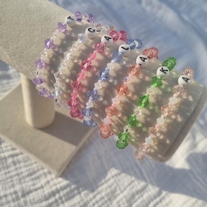 May include: A selection of vibrant beaded bracelets, each with a unique colour palette, including purple, pink, blue, orange, green, and clear beads. White letter beads spell out names on the bracelets, displayed on a neutral-toned surface.