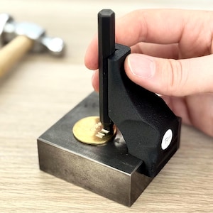 Puede incluir: Una herramienta de estampado de joyas de metal negro con un disco dorado en su lugar. Se inserta un mango negro en la herramienta. Un martillo y otras herramientas están en el fondo. La herramienta está sobre una superficie de madera.