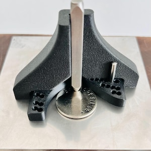 Könnte beinhalten: Ein schwarzes Metallwerkzeug mit einem silbernen Metallstab und einer kreisförmigen Basis. Das Werkzeug hat eine einzigartige Form mit drei ausgestellten Beinen und einem zentralen vertikalen Stab. Die Basis hat eingravierte Zahlen. Das Werkzeug befindet sich auf einer silbernen Metalloberfläche.