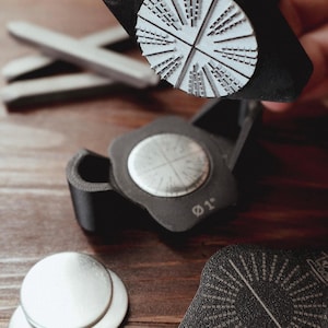 May include: A black stamp tool with a circular stamp head featuring a radial design of architectural elements. The stamp is held by a hand. Additional components include metal discs and a second stamp.