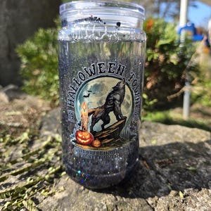 May include: A clear glass tumbler with a lid and straw, filled with black glitter. The tumbler features a Halloween-themed graphic with a howling wolf, a pumpkin, and the words "Halloween Town".