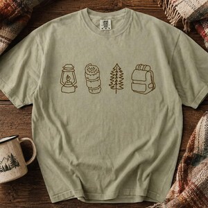 May include: A pale green t-shirt featuring a brown outline of camping symbols: a lantern, food, a fir tree, and a rucksack. A white and black enamel mug with a mountain and tree design is on the left.