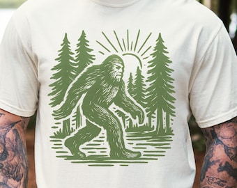 May include: Cream-colored t-shirt featuring a green graphic of Bigfoot walking through a forest with the sun in the background. The design is a detailed illustration with a vintage aesthetic. The shirt is made of a soft, comfortable material.