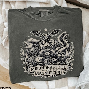 May include: A charcoal gray crewneck t-shirt featuring a cream-colored graphic print of a bat, opossum, raccoon, skunk, and mouse surrounded by stars and foliage. The text MISUNDERSTOOD AND MAGNIFICENT is printed on a banner at the bottom.