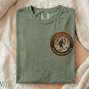 May include: A sage green short-sleeve t-shirt featuring a circular graphic on the chest. The graphic depicts a silhouette of Bigfoot walking through a forest with the text "BELIEVE" at the top and "PACIFIC NORTHWEST FIELD TEAM" at the bottom.