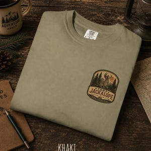 May include: A khaki crew neck t-shirt featuring a graphic on the left chest. The graphic is a badge-shaped illustration of a jackalope in a forest setting with the text "CAMP JACKALOPE" and "WHERE LEGENDS ROAM" printed in a dark brown font.