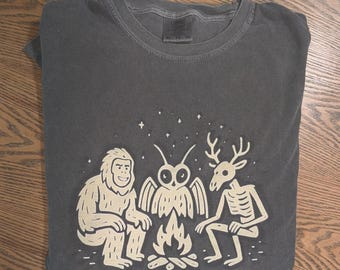 May include: A charcoal gray crew neck t-shirt featuring a cream-colored graphic print of a sasquatch, an owl, and a deer skeleton sitting around a campfire under a starry sky. The shirt has a tag at the neckline indicating size XL.