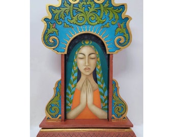 May include: A wooden sculpture of a woman with closed eyes and hands clasped in prayer. She is wearing a blue robe with green floral accents and an orange dress. The sculpture is framed with a gold and blue ornate design and sits on a red base.
