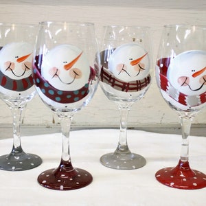 May include: Four wine glasses with snowman faces painted on them. Each snowman has a different colored scarf and the bases of the glasses are painted with polka dots.