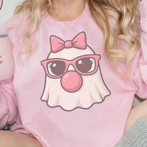May include: Light pink sweatshirt featuring a cartoon ghost wearing pink sunglasses, a bow, and a red nose. The sweatshirt has long sleeves and a crew neck. A pink tumbler is held in the hand. The text "LIGHT PINK" is in a circle.