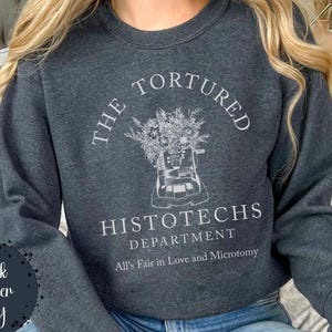 May include: Dark heather gray sweatshirt with the text "THE TORTURED HISTOTECHS DEPARTMENT" and a graphic of a histology machine with flowers. The text "All's Fair in Love and Microtomy" is below the graphic. The words "Dark Heather Gray" are in a circle on the left.