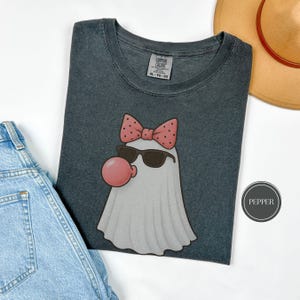 May include: Dark grey t-shirt featuring a cartoon ghost wearing sunglasses and a pink bow with polka dots. The ghost is blowing a pink bubble. The shirt has a Comfort Colors tag and is next to a pair of blue jeans and a tan hat.