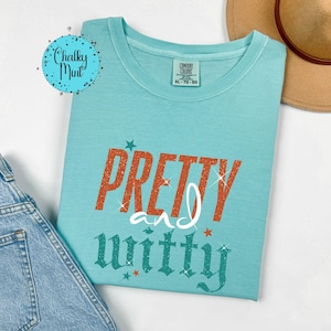 May include: A turquoise Comfort Colors XL-TG-EG t-shirt with the words "PRETTY and witty" in orange glitter and teal lettering. The shirt has a crew neck and short sleeves. A tan hat and denim jeans are in the background.