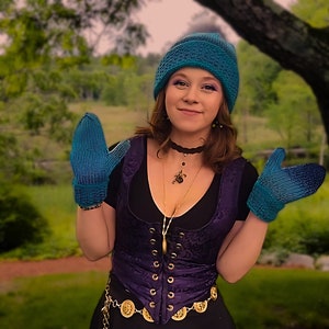 May include: A woman wearing a teal blue knitted hat and matching mittens. She is wearing a purple velvet corset with gold accents and a black top. She is also wearing a gold chain necklace with a pendant.