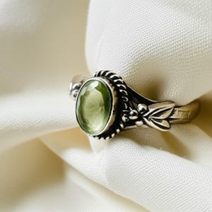 Könnte beinhalten: Ein silberner Ring mit einem ovalen, hellgrünen Edelstein. Der Ring hat ein detailliertes Design mit einem geflochtenen Rand um den Stein und Blattakzenten am Band. Der Ring ist vor einem weißen Hintergrund platziert.