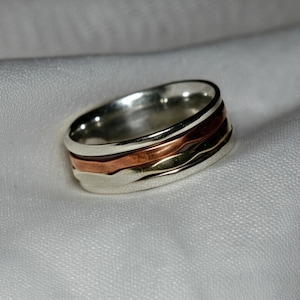 May include: A silver ring with three bands of different metals. The top band is copper, the middle band is silver, and the bottom band is a darker metal. The ring is on a white fabric background.