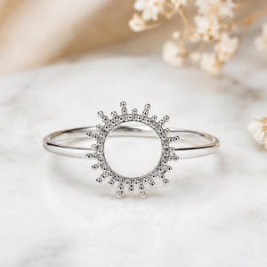 May include: A silver-toned ring featuring a circular centerpiece with a sunburst design. The circular element is adorned with small, bead-like metallic accents radiating outward along the perimeter, attached to a smooth, thin, polished silver-toned band.