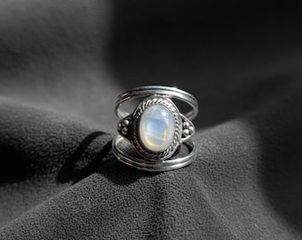May include: A silver ring with an oval, iridescent moonstone center. The ring has a triple-band design with decorative accents on the sides of the moonstone. The ring is set against a dark gray background.