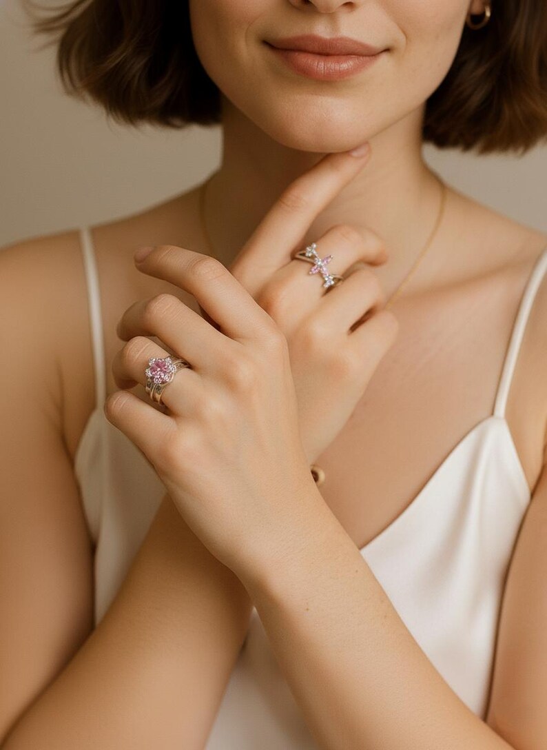 May include: Two silver rings with pink gemstones. One ring features a large, round pink gemstone surrounded by smaller stones. The other ring has a floral design with pink and clear stones. The rings are worn on fingers.