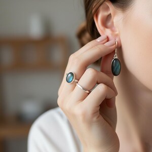 May include: A silver ring featuring an oval labradorite gemstone with blue iridescence, paired with a simple silver band and a matching silver drop earring containing an oval labradorite stone that displays a similar blue iridescent sheen.