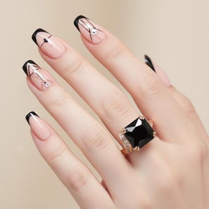 May include: A hand wearing a gold ring with a large, square, black gemstone. The ring is accented with small, clear stones. The nails are painted with a nude base and black tips, with geometric designs.
