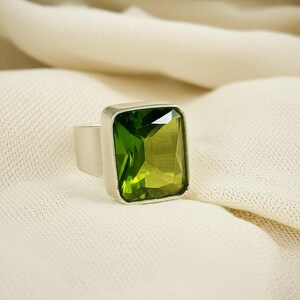 May include: A silver ring featuring a large, rectangular, faceted green gemstone. The gemstone is set in a square bezel, and the band is a simple, polished silver. The ring is displayed on a cream-colored fabric.