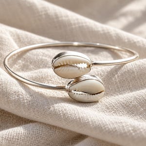 May include: A silver-toned open cuff bracelet featuring two white cowrie shells at the ends. The metal band is smooth and polished, while the shells have a natural, textured appearance with a central slit. The design is simple and circular.