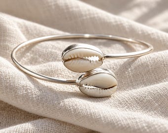 May include: A silver-toned open cuff bracelet featuring two white cowrie shells at the ends. The metal band is smooth and polished, while the shells have a natural, textured appearance with a central slit. The design is simple and circular.