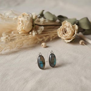May include: A pair of oval-shaped earrings featuring polished labradorite gemstones set in silver-toned metal. Each earring has a curved hook wire for attachment, showcasing the natural grey and iridescent blue flashes within the stone surface.