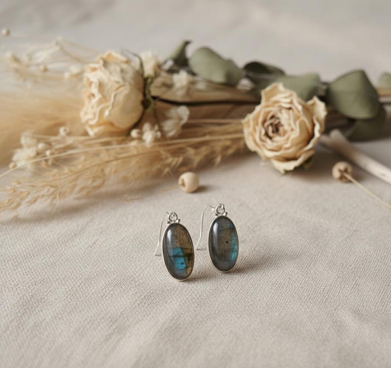 May include: A pair of oval-shaped earrings featuring polished labradorite gemstones set in silver-toned metal. Each earring has a curved hook wire for attachment, showcasing the natural grey and iridescent blue flashes within the stone surface.