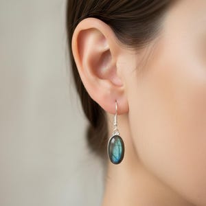 May include: A silver drop earring featuring an oval-shaped labradorite gemstone. The stone displays a dark, iridescent blue and grey surface with natural inclusions. The earring hangs from a simple silver hook wire, showcasing a polished finish.